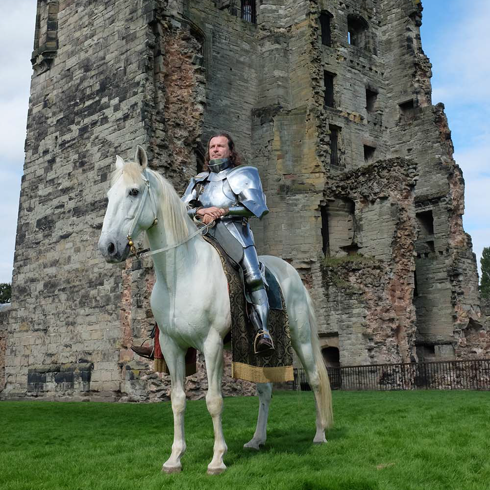 Jason sits on the back of a beautiful white horse. Behind him looms the remains of a grade castle.