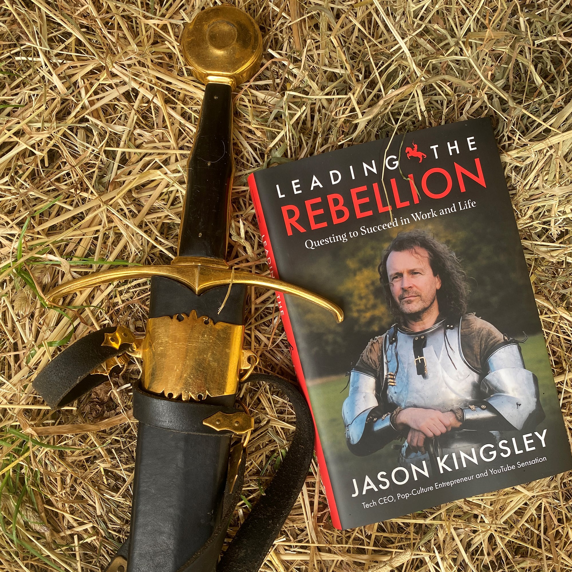 Jason's book Leading the Rebellion sits on some hay next to an authentic sword with gold detailing. Jason's upper body adorns the cover of his book, he's wearing Medieval armour.