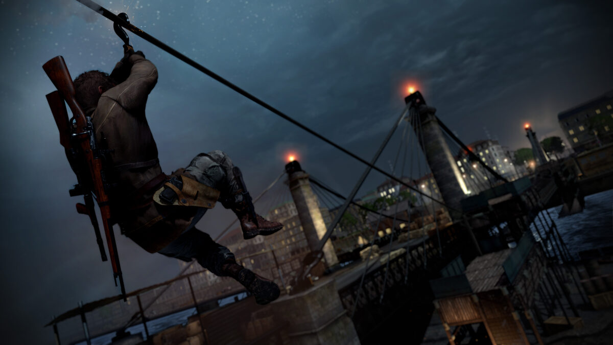 Harry Hawker slides down a zipline toward a wire bridge made out of steel and concrete. Lights illuminate from the top of the pillars on the bridge. A town can be seen in the distance.